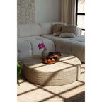 Pouf rond en jonc de mer tressé naturel sur un tapis rond devant un canapé écru avec des pâtisseries et un vase