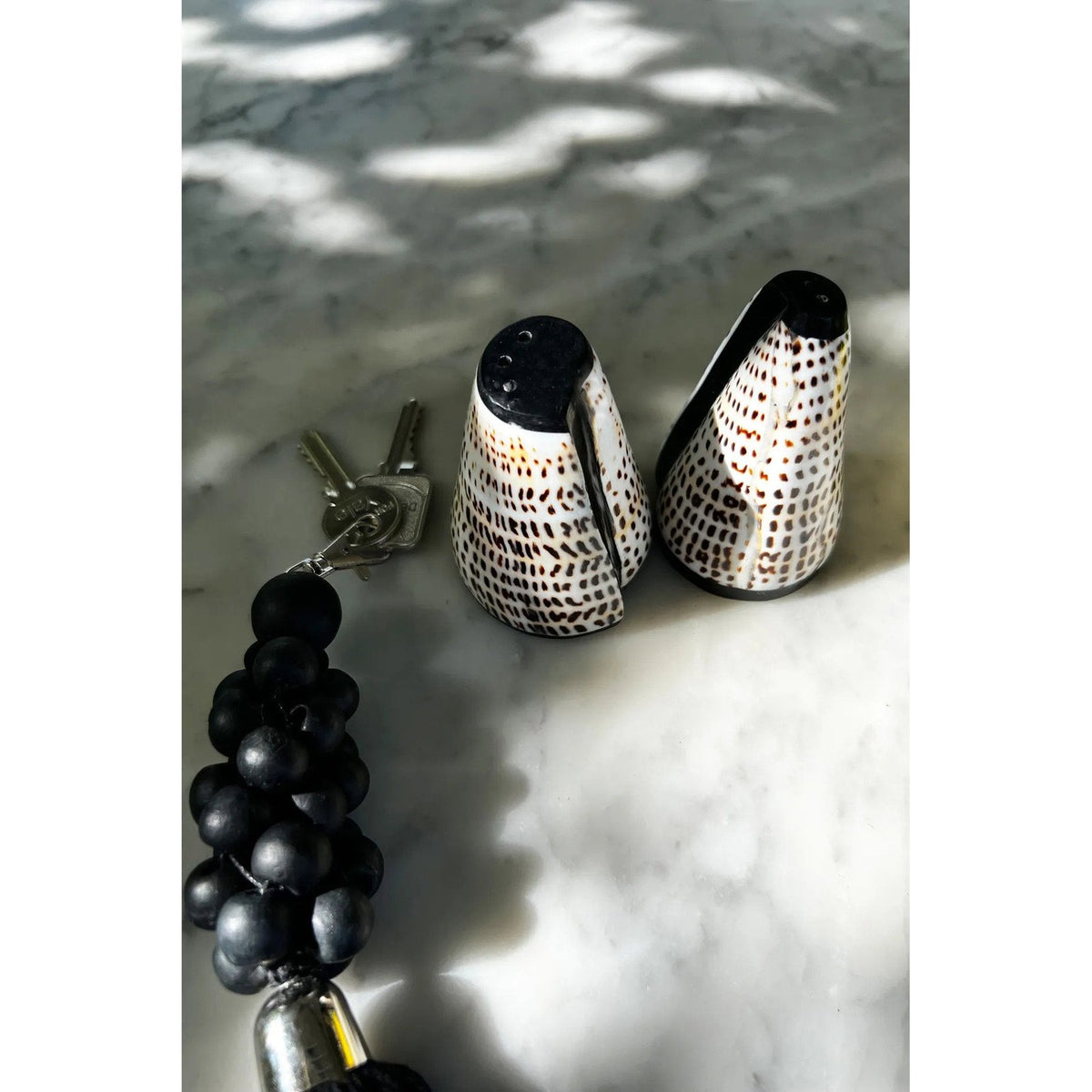 Salière et poivrière en coquillage posées sur un marbre à côté d'un trousseau de clés à perles noires