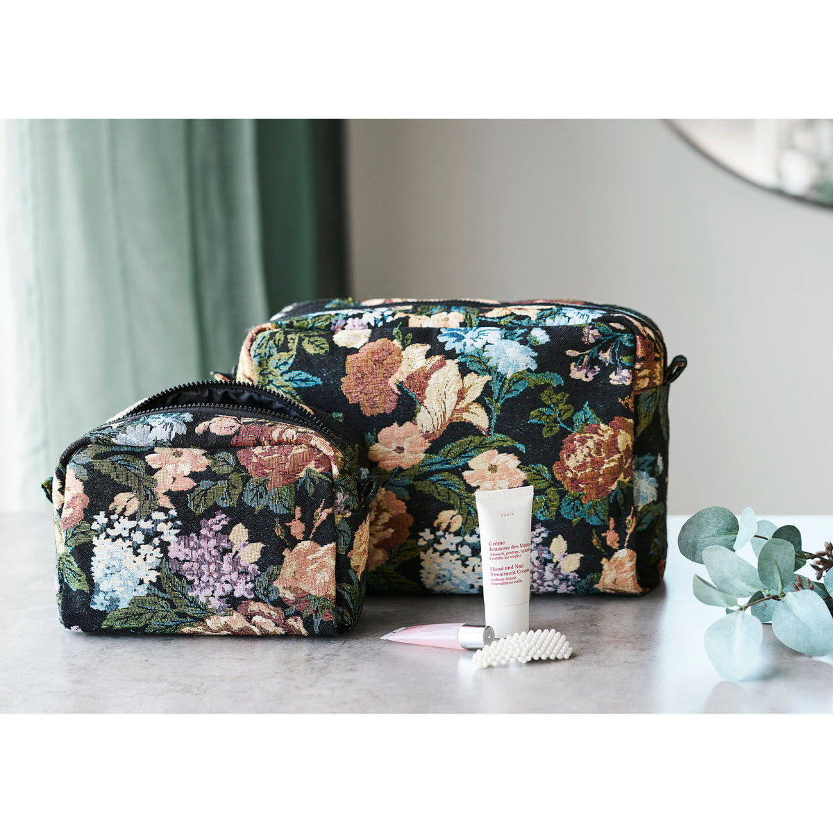 Les deux trousses Zoe posées sur un plan minéral avec une crème pour les mains et une branche d'eucalyptus
