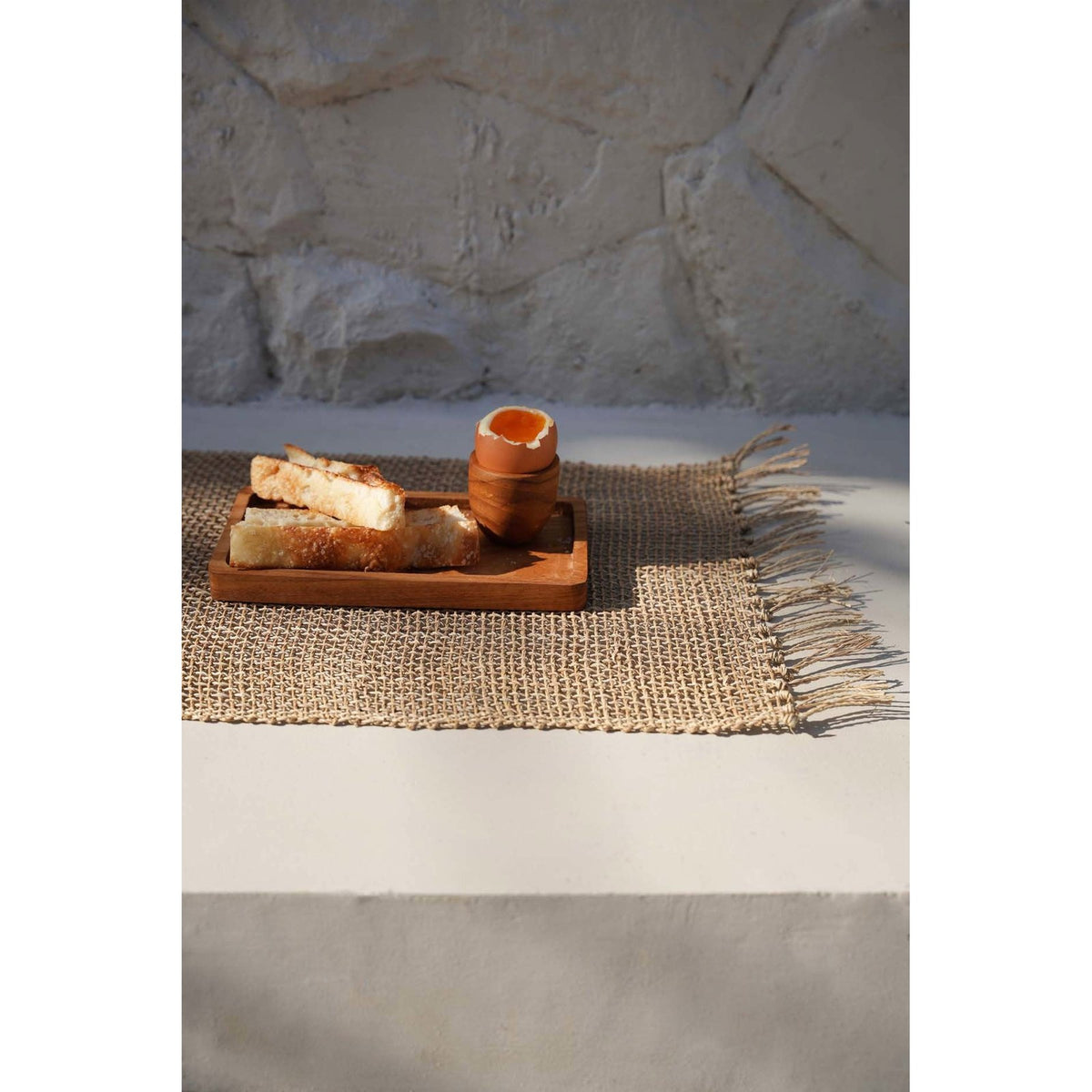 Set de table Isey posé sur un banc de pierre au soleil avec un plateau en teck et un oeuf à la coque