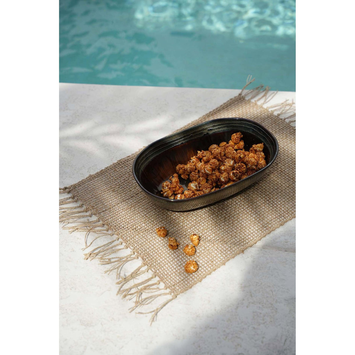 Set de table Isey au bord d'une piscine avec un plat ovale sombre et du pop-corn caramélisé