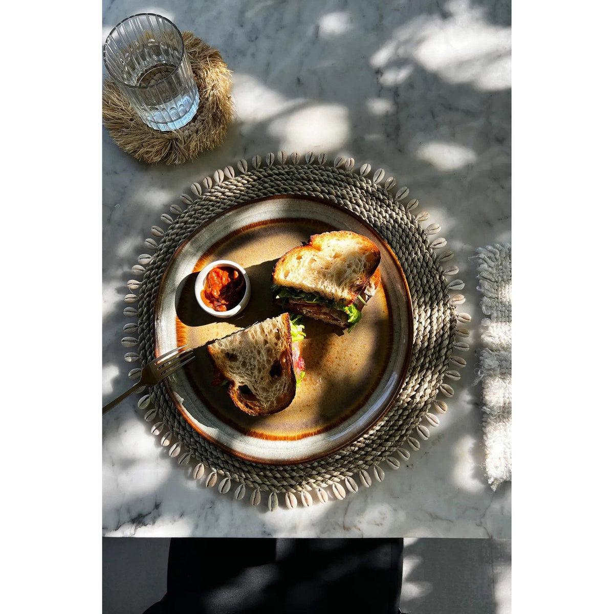 Set de table en jonc de mer naturel bordé de cauris sous une assiette à bord émaillé vu de près