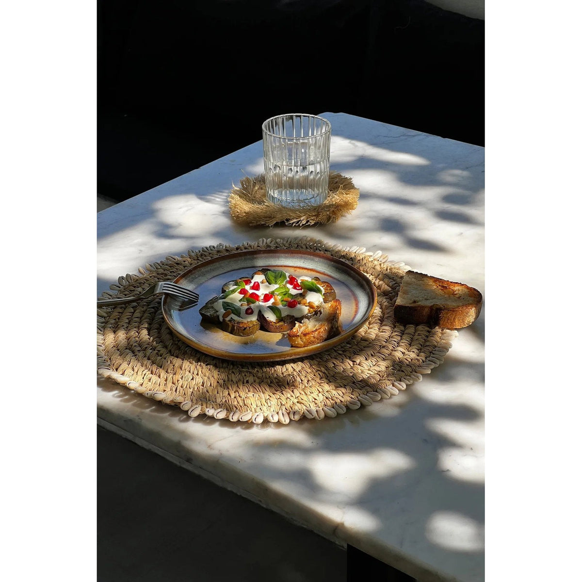 Set de table rond en raphia et cauris sous une assiette émaillée avec du pain sur une table en marbre près d'un verre