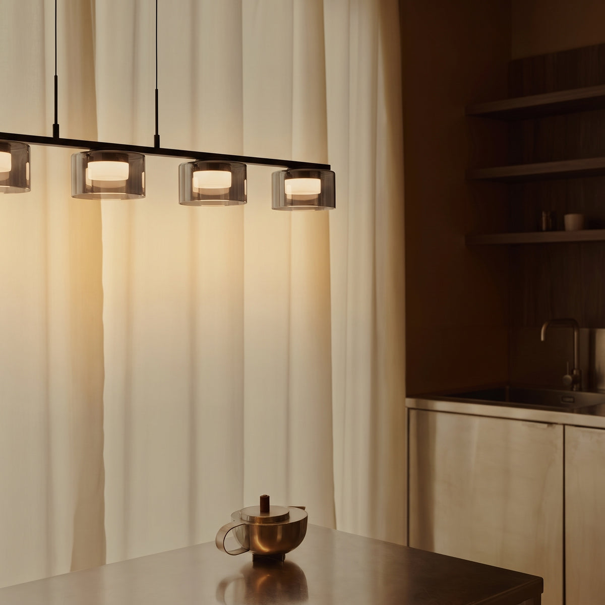 Suspension Cholet rampe LED allumée au-dessus d'un îlot de cuisine en inox, devant un rideau crème