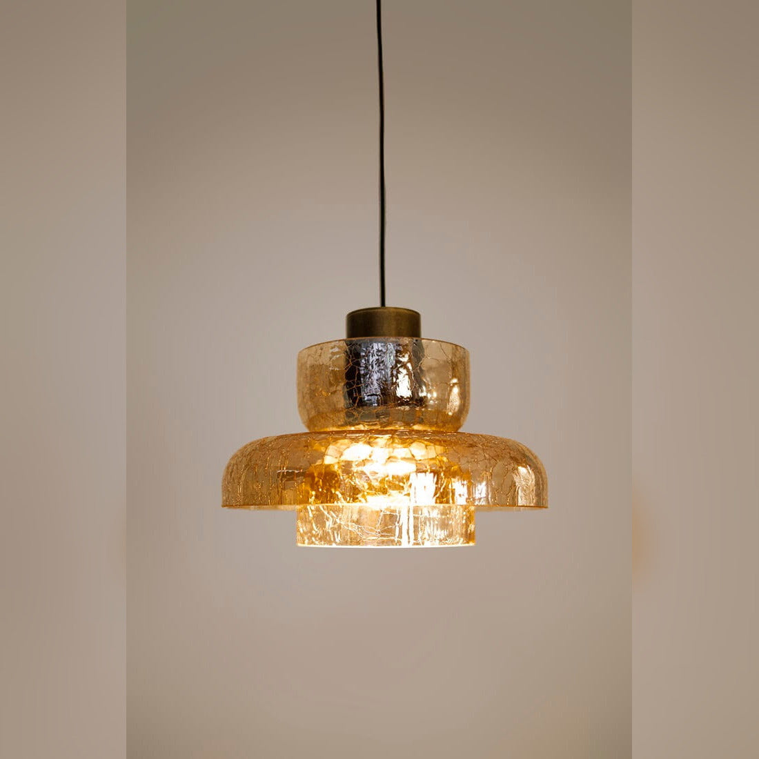 Suspension Signature C allumée sur un mur beige verre ambré craquelé traversé par la lumière