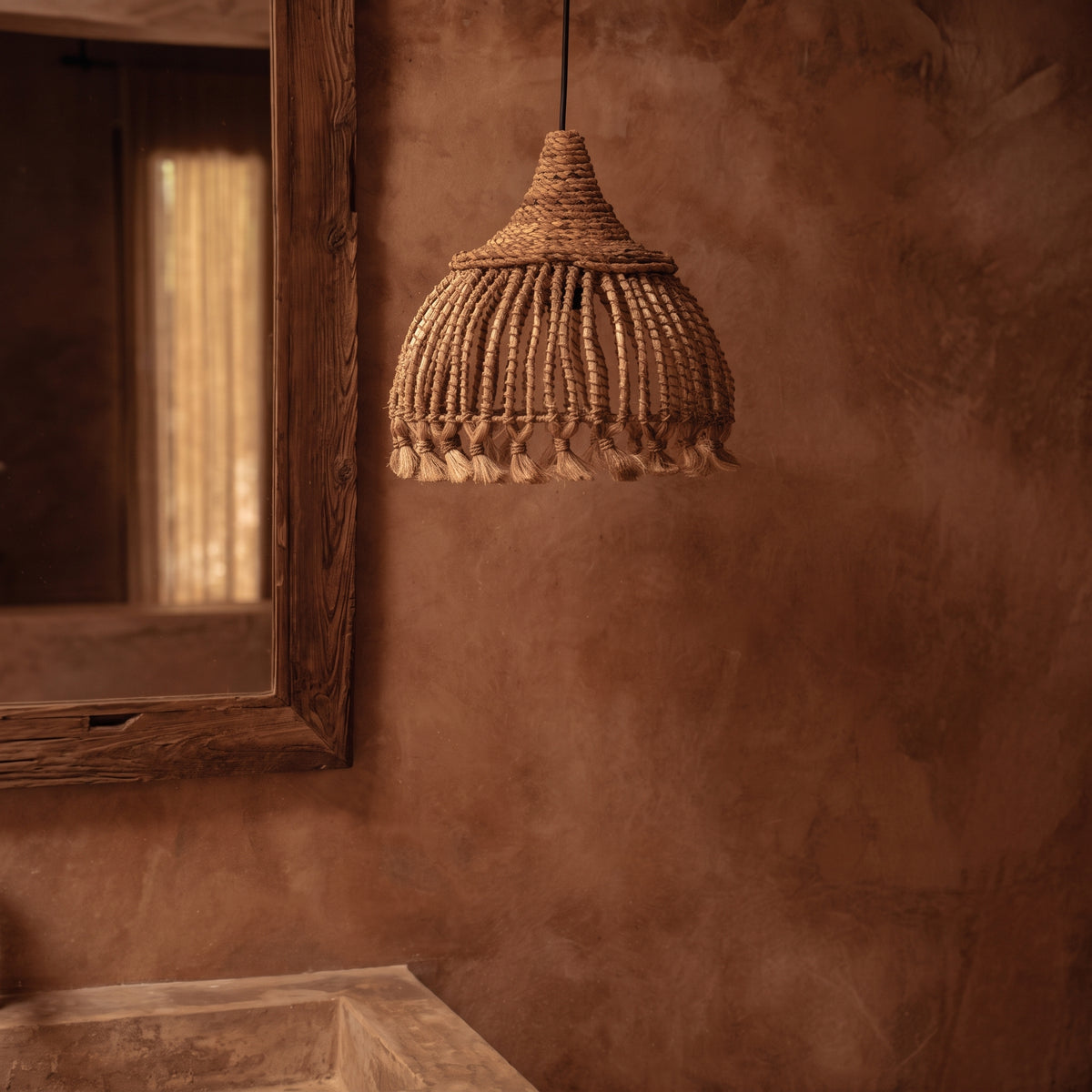 Suspension Tassel en abaca naturel devant un mur terracotta, à côté d'un miroir au cadre en bois