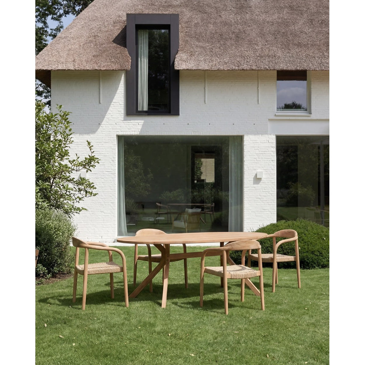 Table à manger ovale Kulala entourée de quatre fauteuils à assise tressée devant une maison blanche