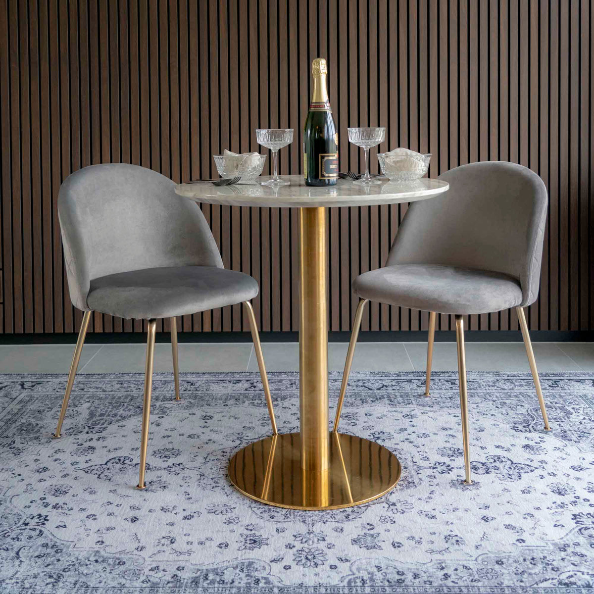 Table Bolzano dressée avec une bouteille et des coupes entre deux chaises en velours gris