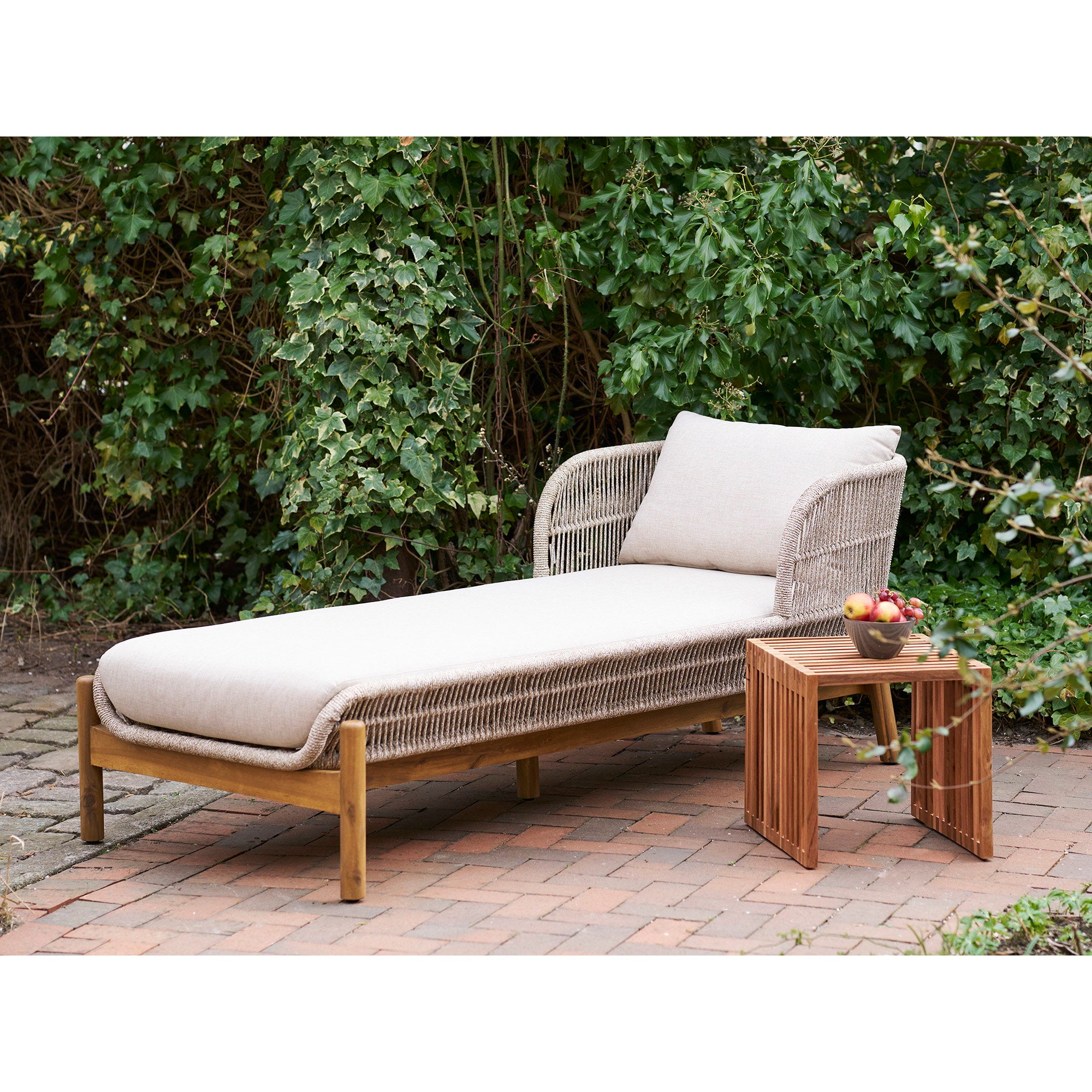 Table d'appoint Pego teck naturel lames verticales posée sur terrasse en brique avec chaise longue