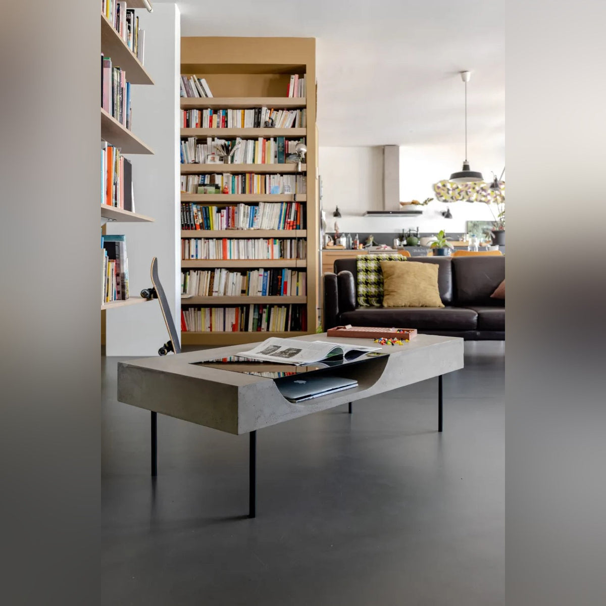 Table basse Curb en béton dans un salon avec un magazine ouvert un ordinateur portable et une bibliothèque en fond