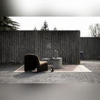 Table basse Strut carrée sur un tapis en plein air à côté d'un fauteuil marron devant un mur en béton strié