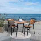 Table bistro Cleveland en teck et métal noir sur une terrasse face à la mer avec deux chaises à lattes et une lanterne boule