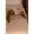 Table basse Ashley Cocoon en teck sur un lit aux draps clairs avec un ordinateur portable ouvert