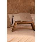 Table basse Ashley Cocoon en teck posée sur un lit avec un ordinateur portable devant un mur enduit