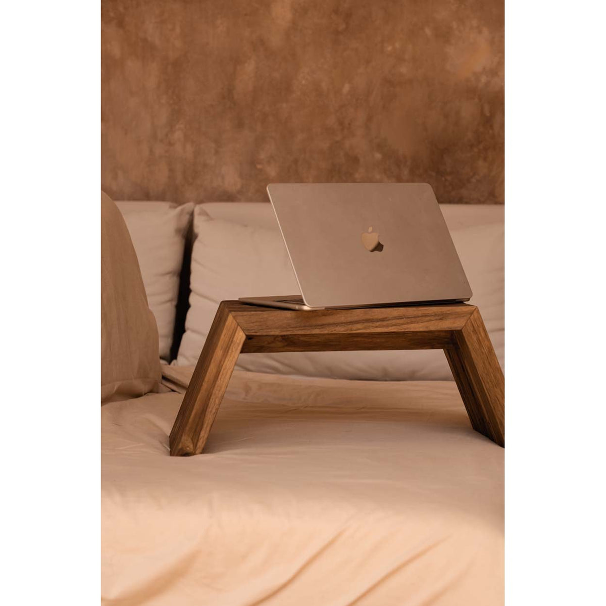 Table basse Ashley Cocoon en teck posée sur un lit avec un ordinateur portable devant un mur enduit