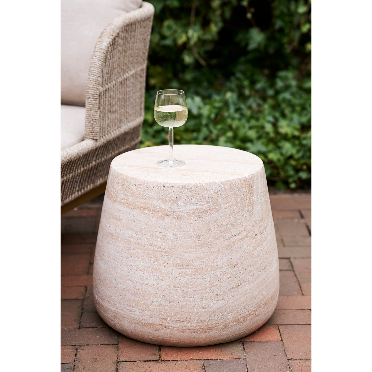 Table d'appoint Bonn effet travertin sur une terrasse en brique avec un verre de vin blanc près d'un fauteuil tressé
