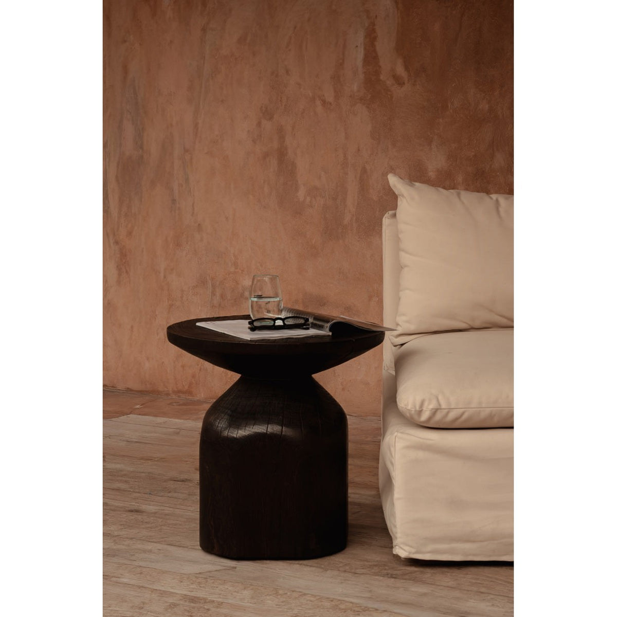 Table d'appoint Drumline teinte noyer foncé avec un verre d'eau et un magazine ouvert à côté d'une chauffeuse écrue