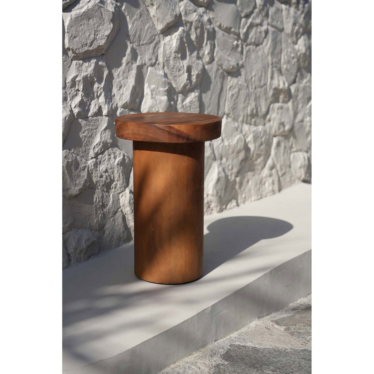 Table d'appoint Inti en bois posée au soleil devant un mur de pierre blanche