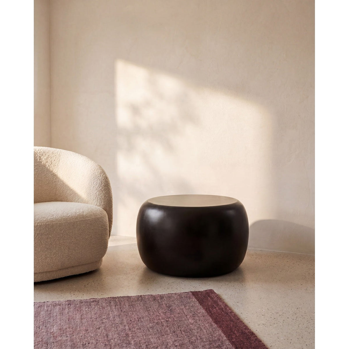 Table d'appoint ronde Matt en résine noire posée sur un sol en terrazzo près d'un fauteuil bouclé écru