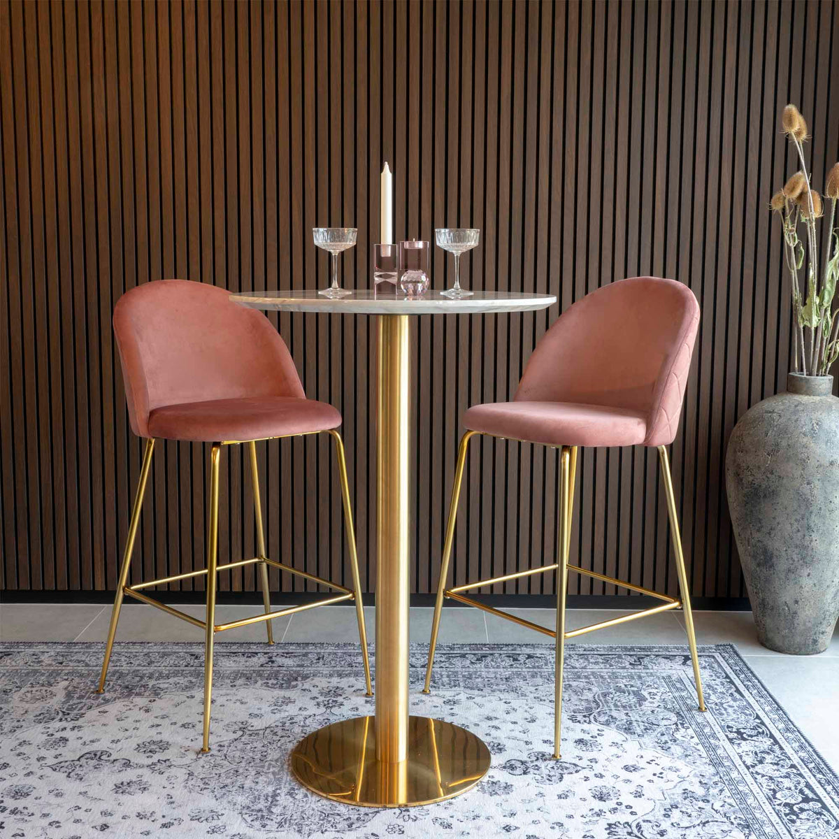 Table de bar Bolzano entre deux tabourets en velours rose sur un tapis devant un mur à tasseaux