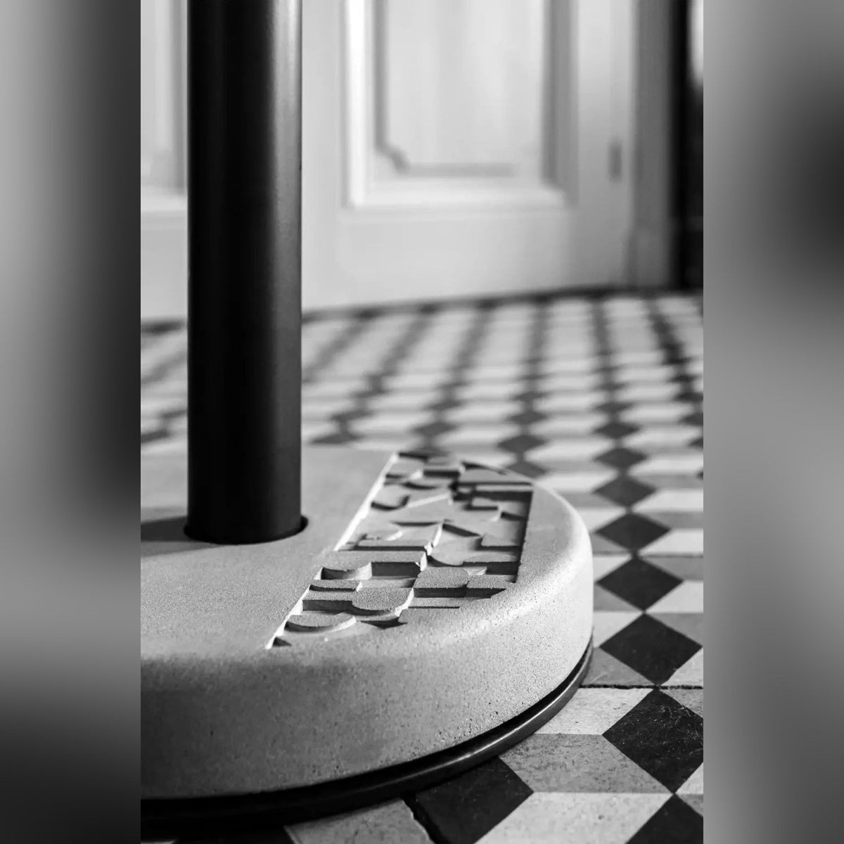 Socle en béton à frise en relief et colonne noire de la table Donut sur un carrelage en damier noir et blanc