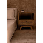 Table de chevet Breeze en teck et rotin au bord d'un lit aux draps beiges contre un mur enduit