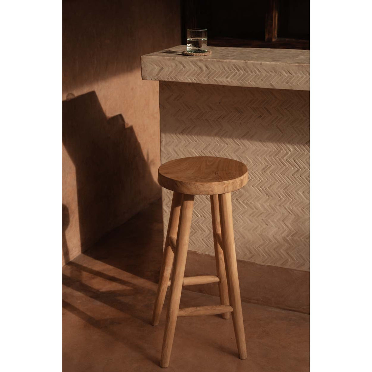 Tabouret de bar Kedut en teck devant un comptoir à motif en chevrons avec un verre posé dessus