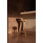 Tabouret de bar Kedut en teck près d'un comptoir dans la lumière rasante d'un mur en enduit terracotta