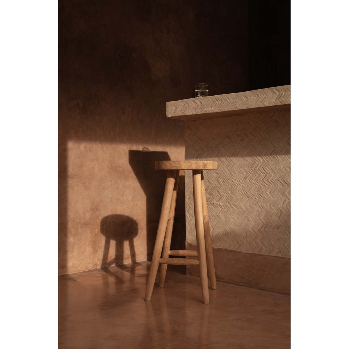 Tabouret de bar Kedut en teck près d'un comptoir dans la lumière rasante d'un mur en enduit terracotta