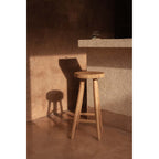 Tabouret de bar Kedut en teck au pied d'un comptoir en enduit avec son ombre portée sur le mur