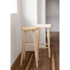 Tabouret de bar Kedut en teck devant un autre tabouret à assise carrée dans une pièce blanche au parquet foncé
