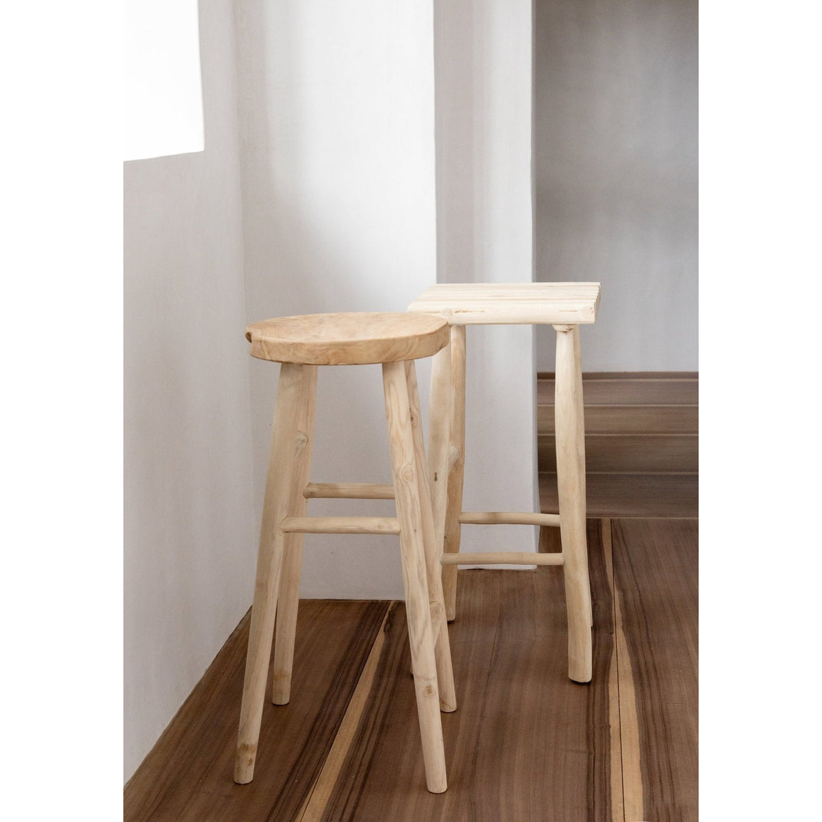 Tabouret de bar Kedut en teck devant un autre tabouret à assise carrée dans une pièce blanche au parquet foncé