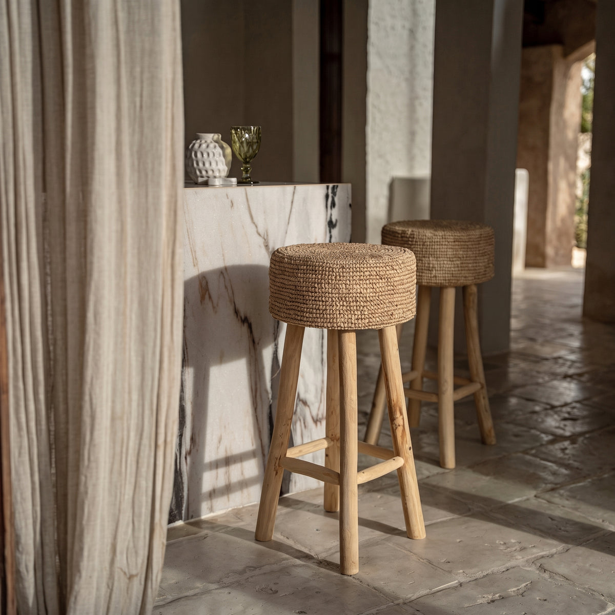 Deux tabourets de bar Raffia le long d'un bar en marbre veiné, près d'un voilage en lin