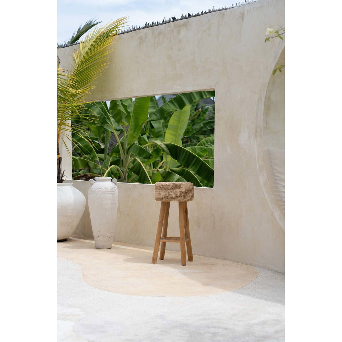 Tabouret de bar Raffia en teck et raphia devant un mur blanc ouvert sur des bananiers près de deux jarres