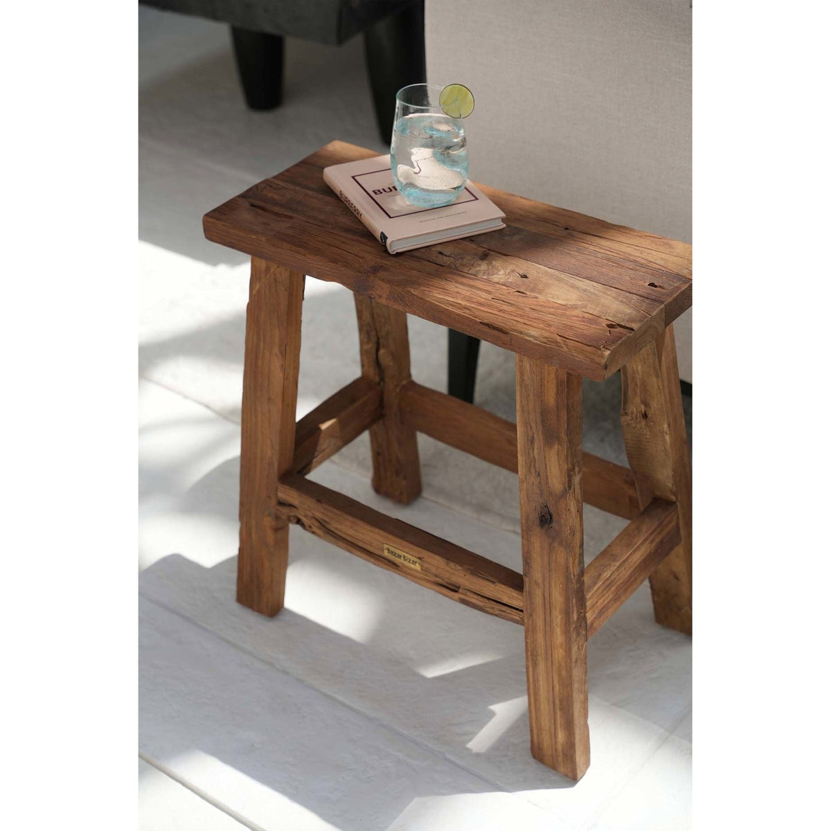 Tabouret rustique Fab en teck portant un verre et un livre au bout d'un canapé