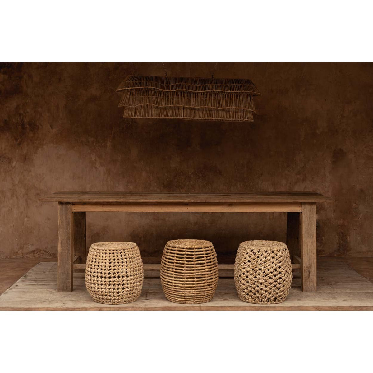 Tabouret Gustih au centre de trois tabourets tressés sous une longue table en bois brut et une suspension en fibre