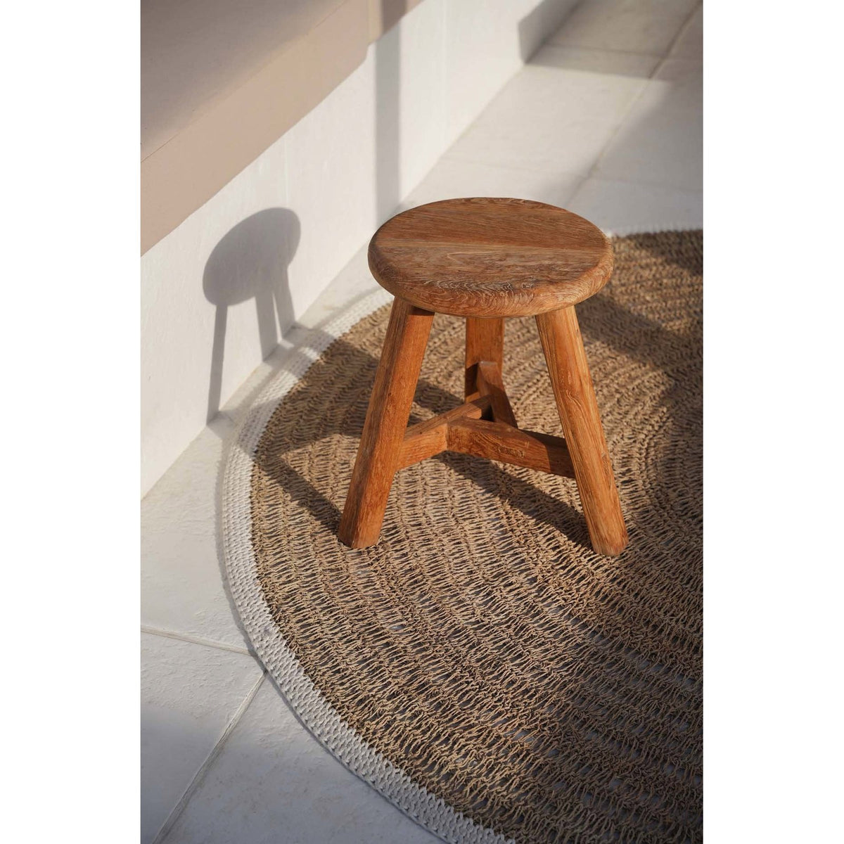 Tabouret Noguchi en teck posé sur un tapis rond tressé au soleil près d'une banquette blanche
