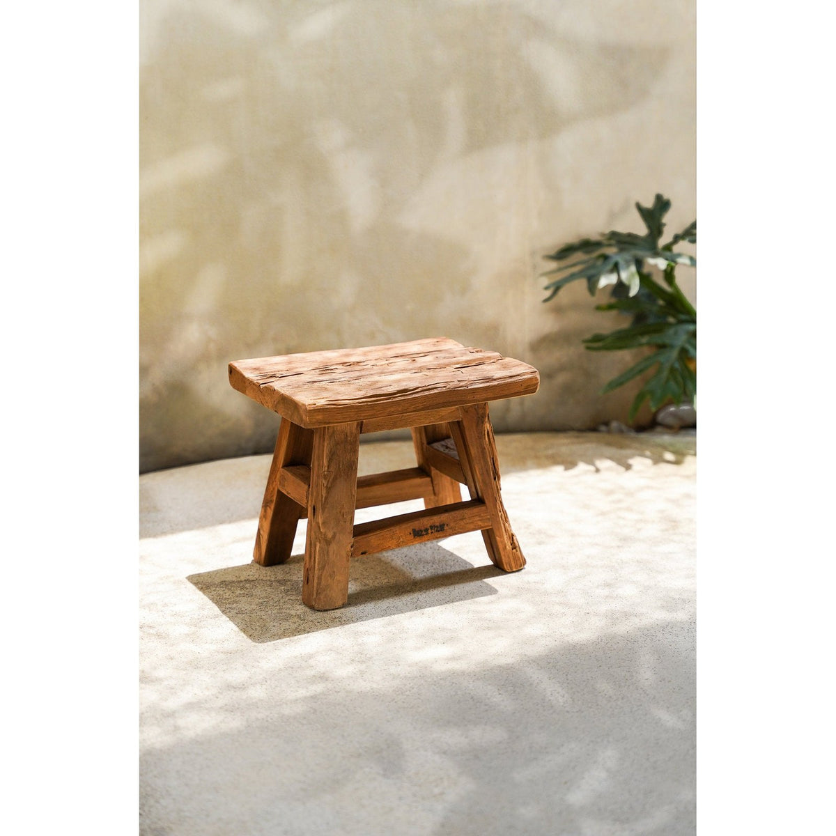 Tabouret Yoke petit modèle en teck recyclé posé au soleil devant un mur enduit et une plante