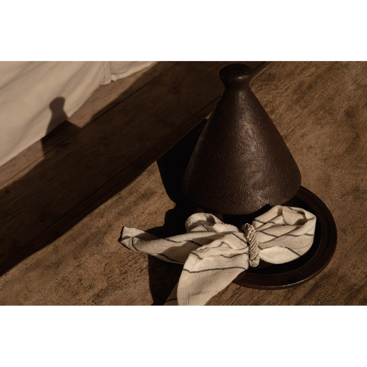 Tajine brun foncé sur une table en bois avec une serviette en lin rayé et un rond de serviette torsadé