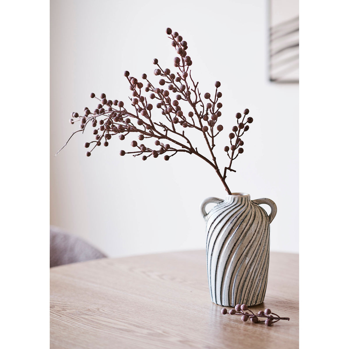 Vase amphore rayé bleu garni d'une branche de baies sur une table en bois clair