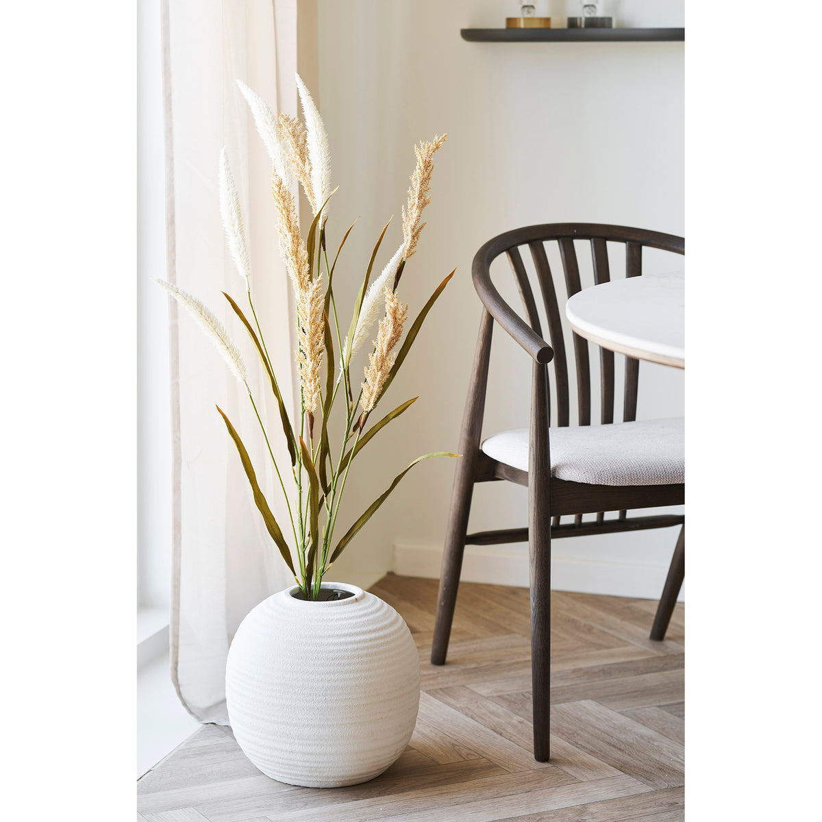 Vase boule en céramique blanche garni de graminées posé au sol à côté d'une chaise en bois foncé