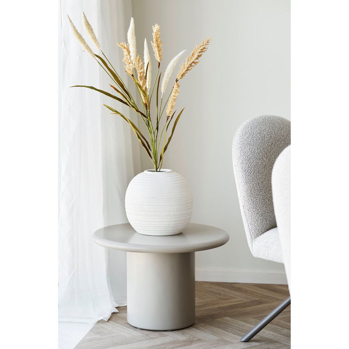 Vase boule en céramique blanche garni de graminées sur une table d'appoint grise près d'un fauteuil bouclé