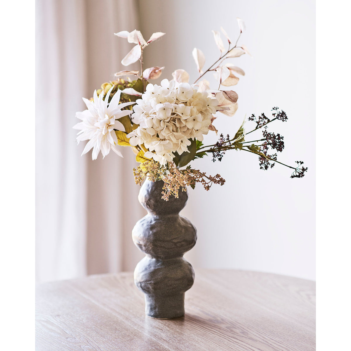 Vase gris taupe moucheté garni d'un bouquet blanc de dahlias et d'hortensias sur une table en bois