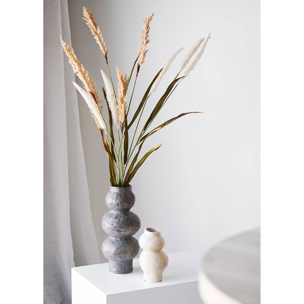 Vase gris taupe moucheté garni de hautes graminées sur un socle blanc à côté d'un petit vase crème