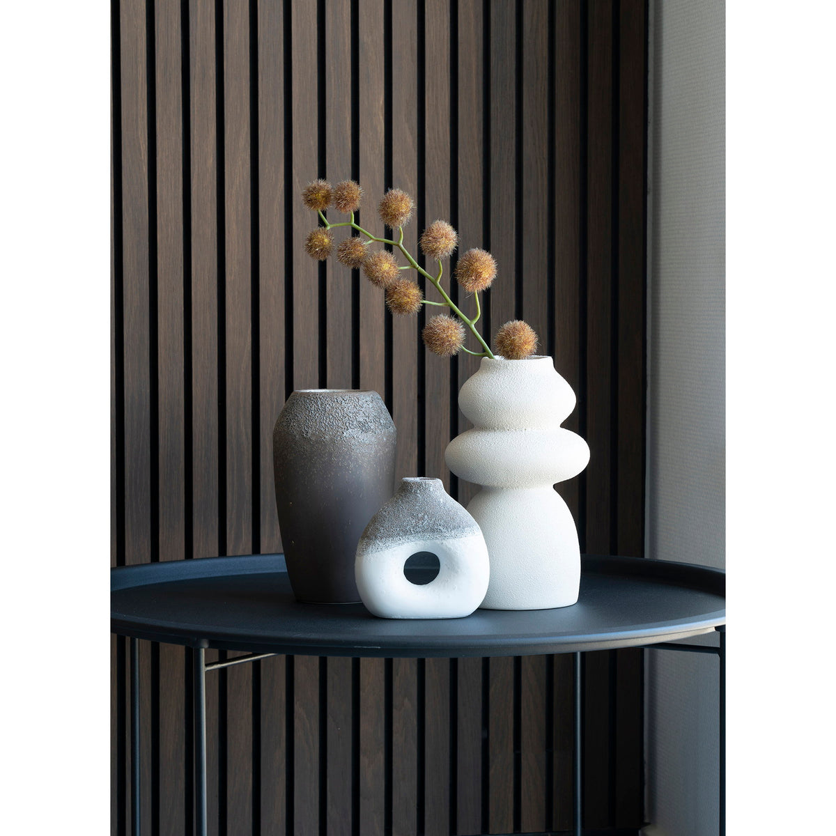 Vase totem en grès beige avec deux autres vases sur une table ronde noire devant un mur à lattes
